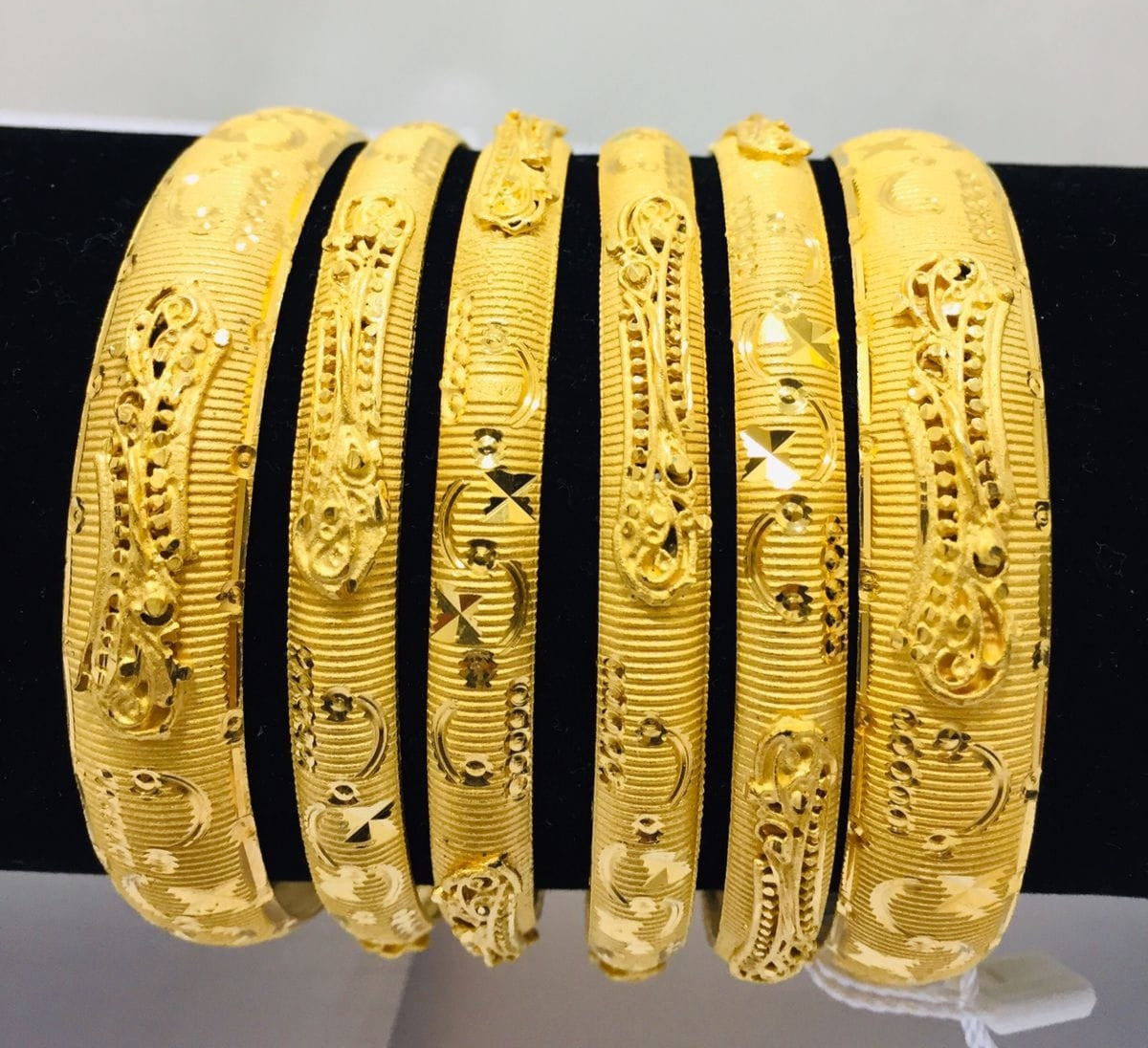 A set of gold-colored metal bangles with intricate filigree work, displayed against a black background.